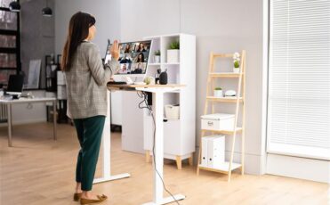A woman employee standing at an adjustable-height desk and waving to colleagues on a computer screen during a remote video call,